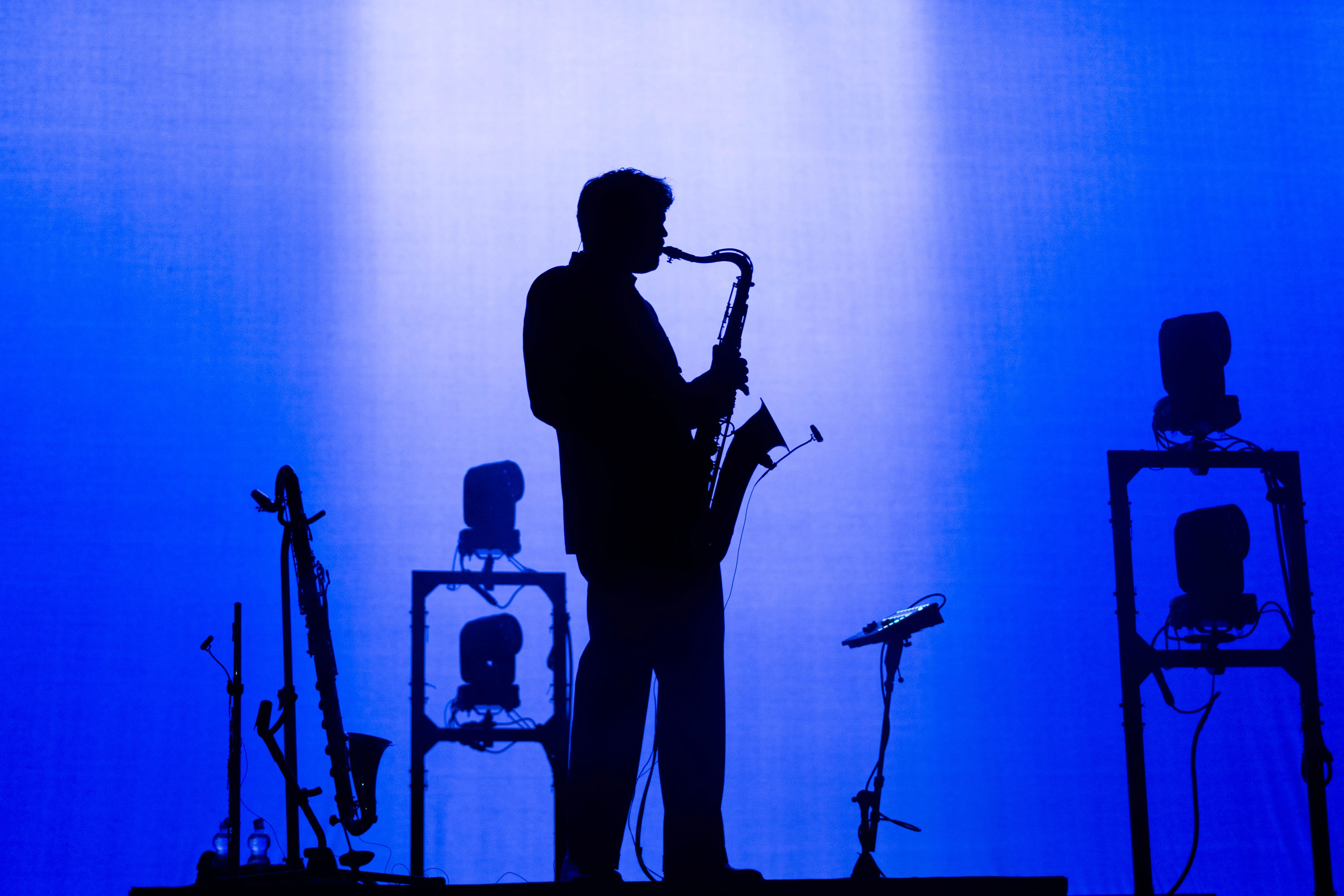 Een silhouet van een muzikant die een saxofoon speelt tegen een blauwe achtergrond met lichtopstellingen.