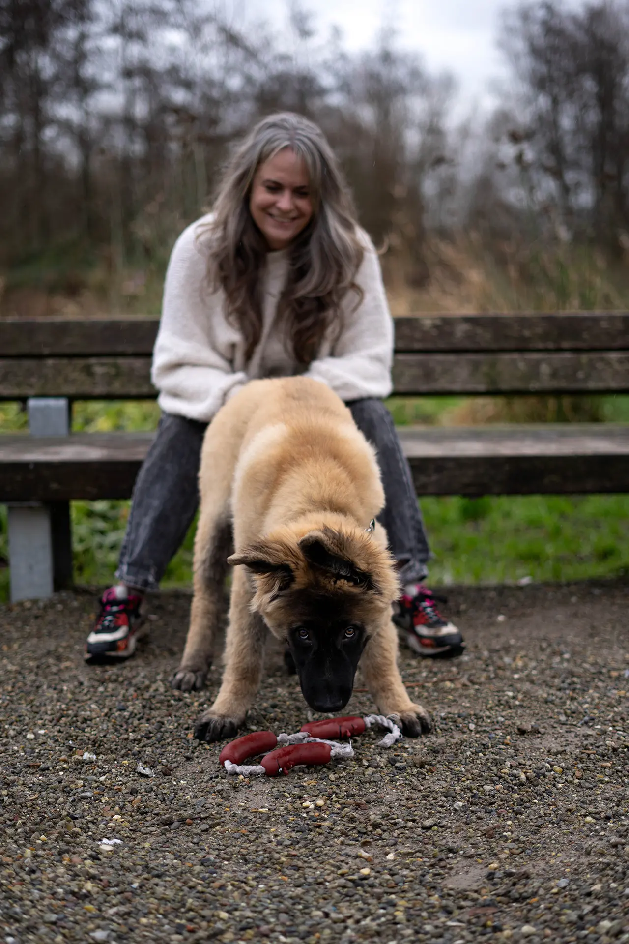 Een vrouw zit op een bank en kijkt lachend naar een puppy die interesse heeft in een speeltje op de grond.
