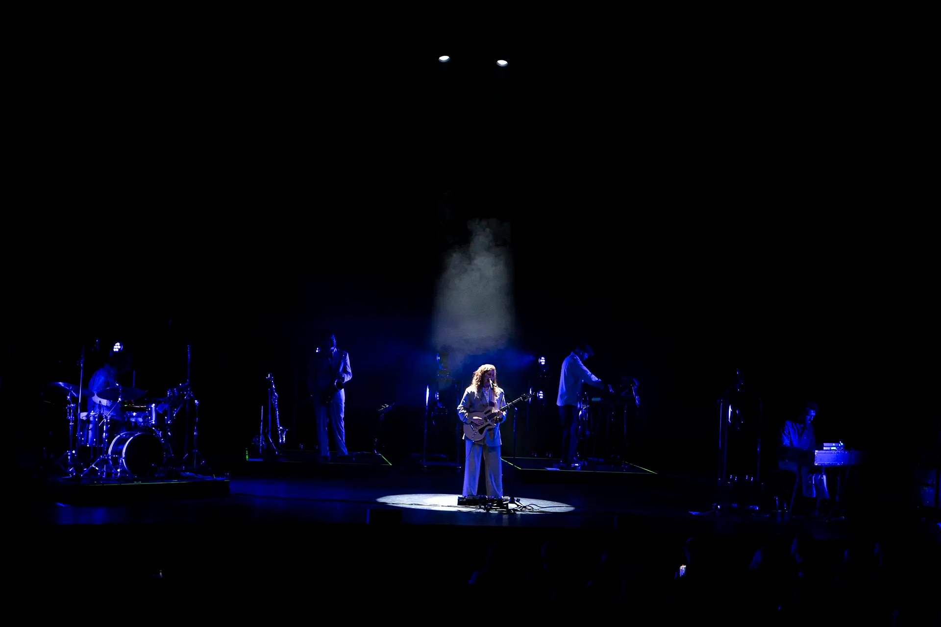 Een muzikante met een gitaar staat in het middelpunt van een podium, omringd door bandleden en subtiel blauw licht.
