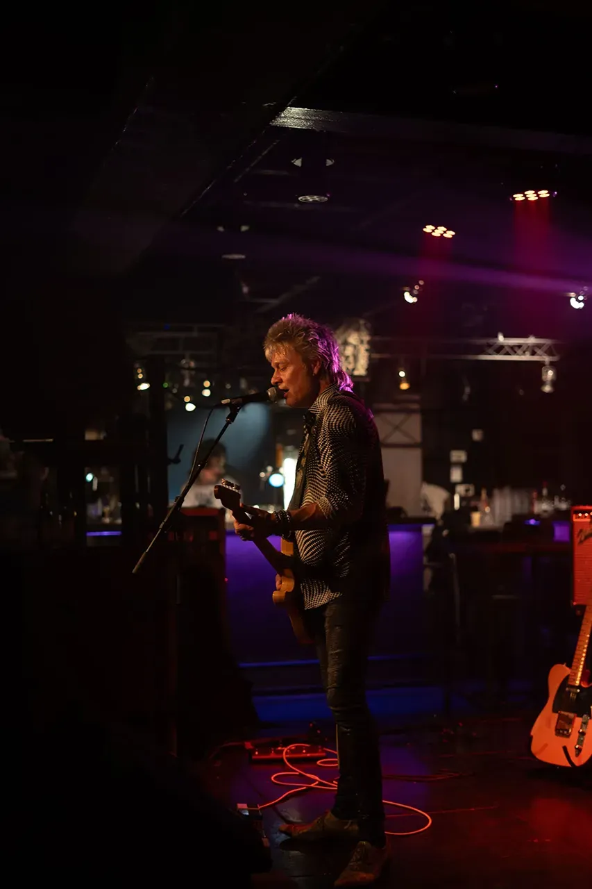 Een gitarist zingt en speelt op een podium in een sfeervolle, lichtgevende muziekclub.