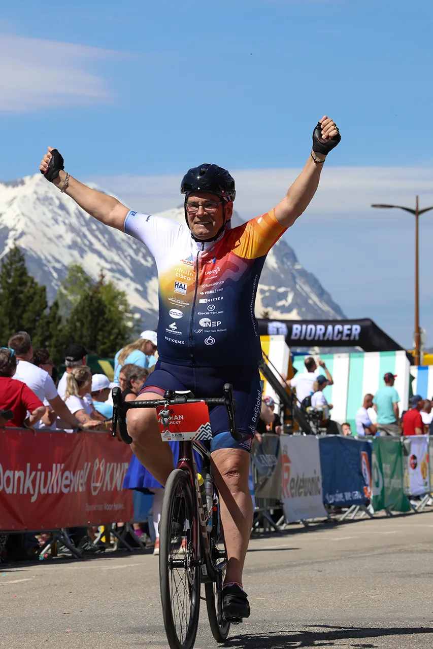 Een jubelende wielrenner steekt zijn armen omhoog in de lucht na het voltooien van een race, met bergen op de achtergrond.